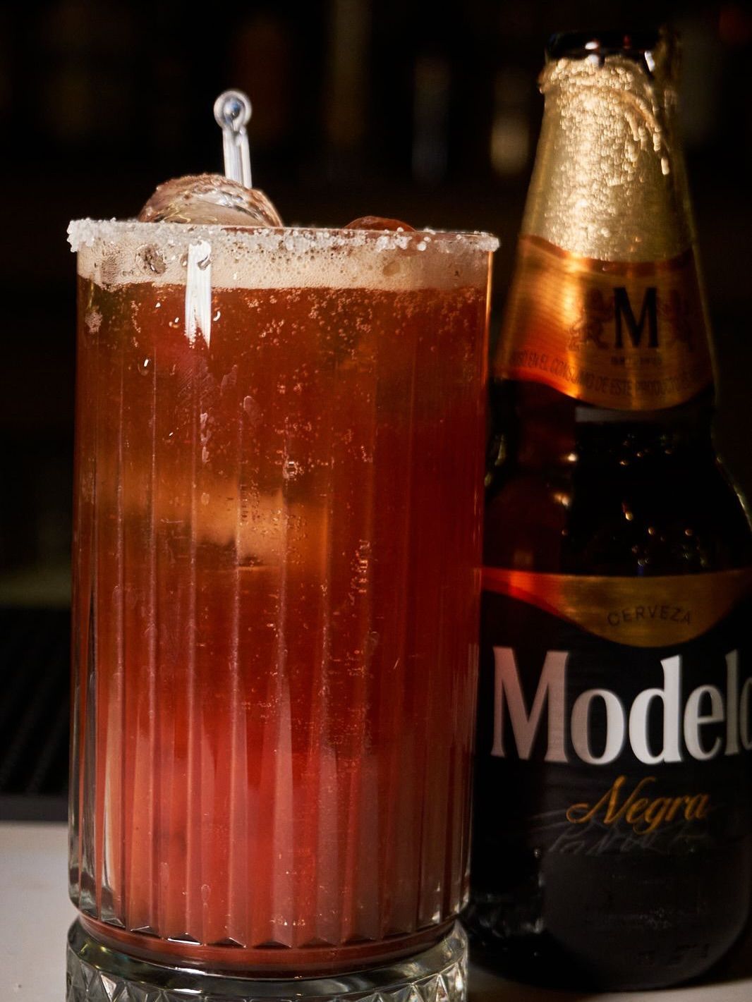 Un cóctel rojo alto con pajita junto a una botella de cerveza Modelo sobre una mesa oscura.