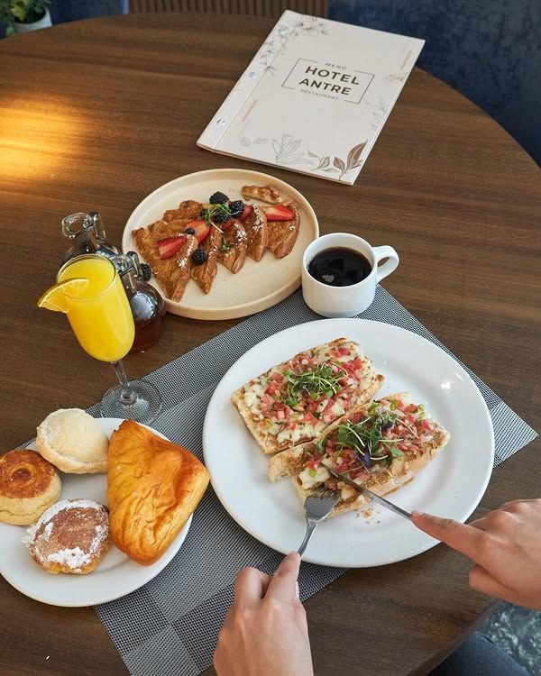 El desayuno servido sobre una mesa: pasteles, fruta, café, zumo de naranja y manos cortando tostadas como si fueran pizza.