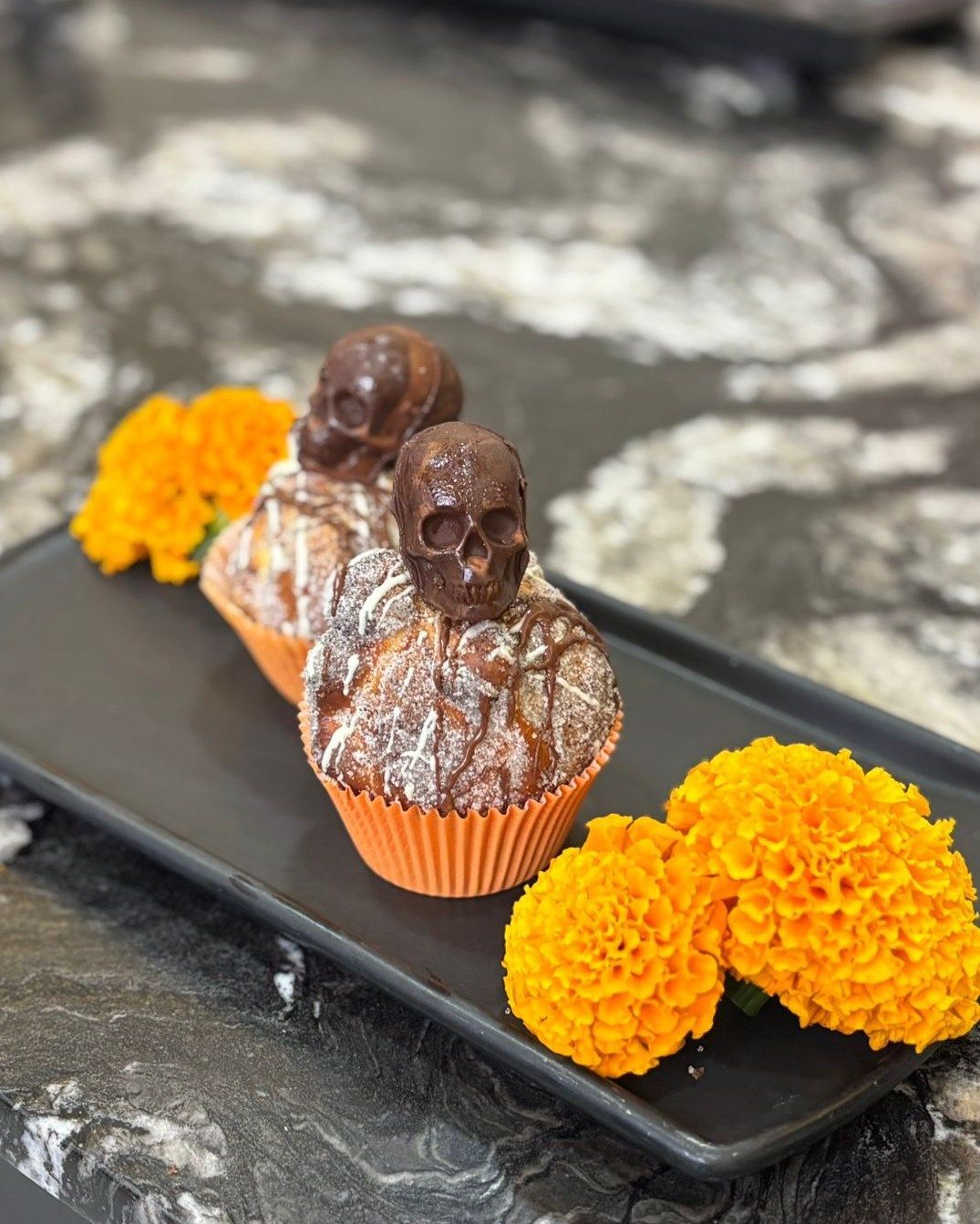Tres pasteles cubiertos de chocolate en una bandeja negra con flores de color naranja brillante sobre una encimera oscura.