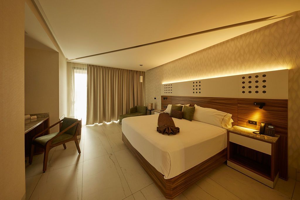Cama bien hecha con ropa de cama gris, almohadas verdes y cabecero de madera en una habitación de hotel moderna.