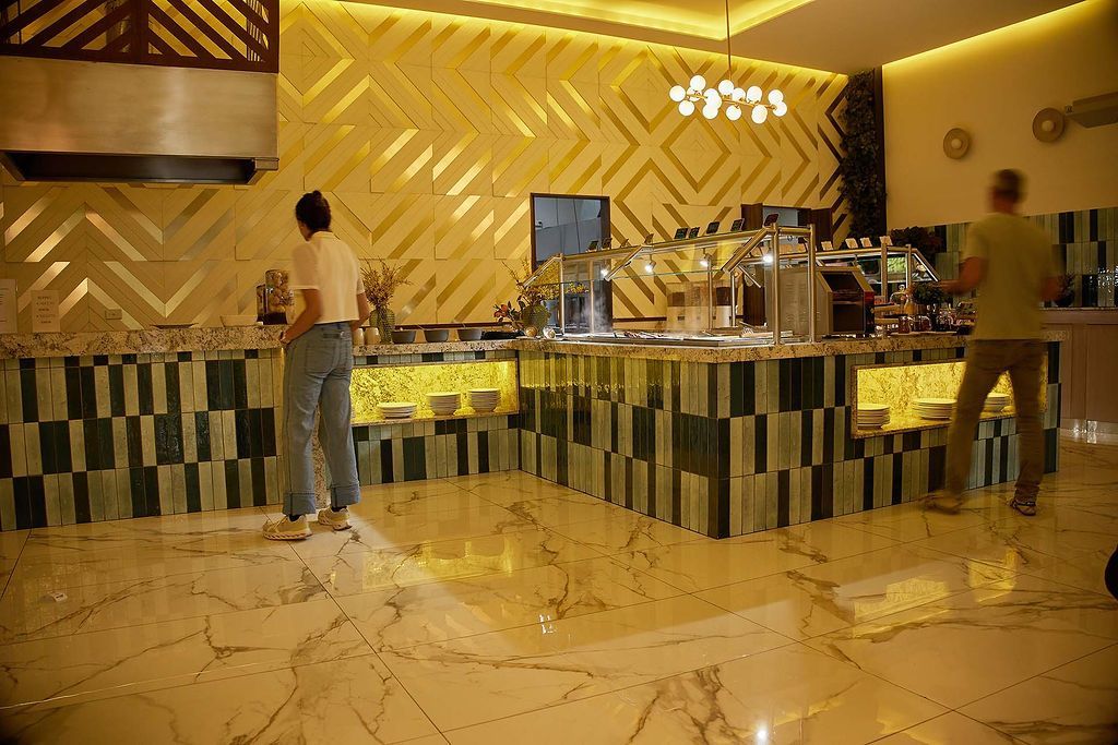 Mostrador de buffet de hotel moderno con dos personas sirviendo comida bajo una iluminación amarilla cálida.