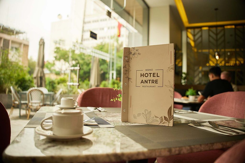 Menú del hotel sobre una mesa de cafetería con una taza y un platillo blancos, en un luminoso patio al aire libre.