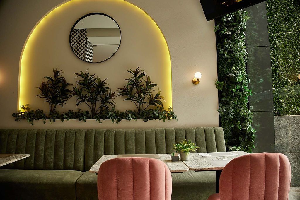 Acogedor reservado de estilo moderno con asientos tapizados en verde, sillas rosas, espejo redondo e iluminación cálida.