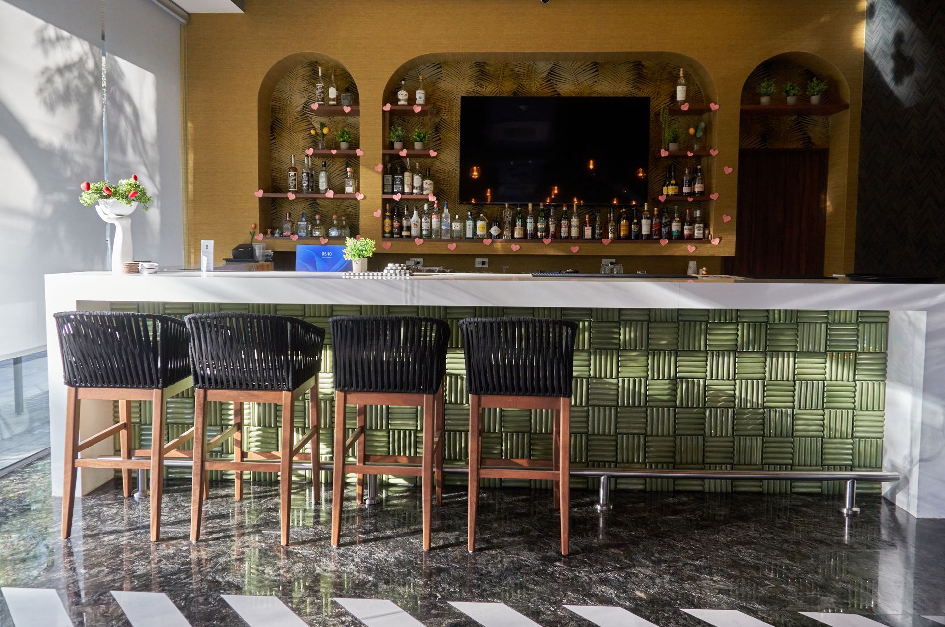 Bar moderno con barra de azulejos verdes, taburetes negros y un televisor montado en la pared detrás de la barra.