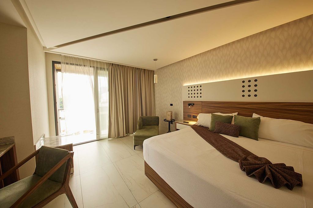 Habitación de hotel moderna con una cama grande, puertas que dan al balcón, cortinas y una cálida decoración en tonos beige.