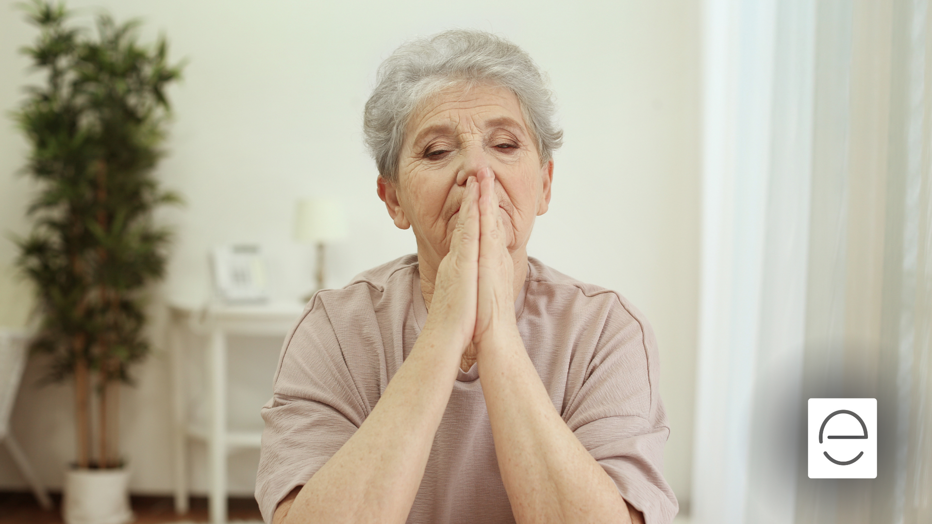 An elderly woman is praying with her hands folded in front of her face.