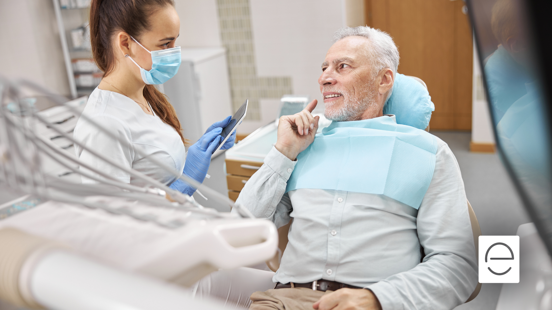Dentist in mask showing older patient tablet. Dental tools, chair, and light present.