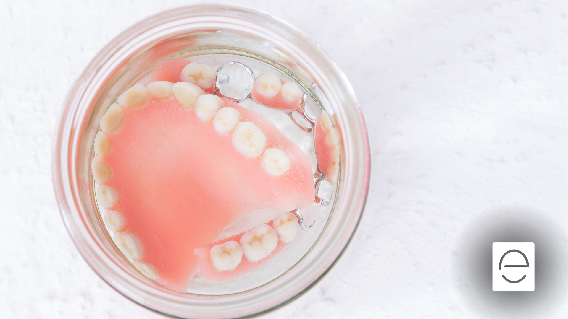 Dentures submerged in clear liquid within a glass jar, resting on a white surface.