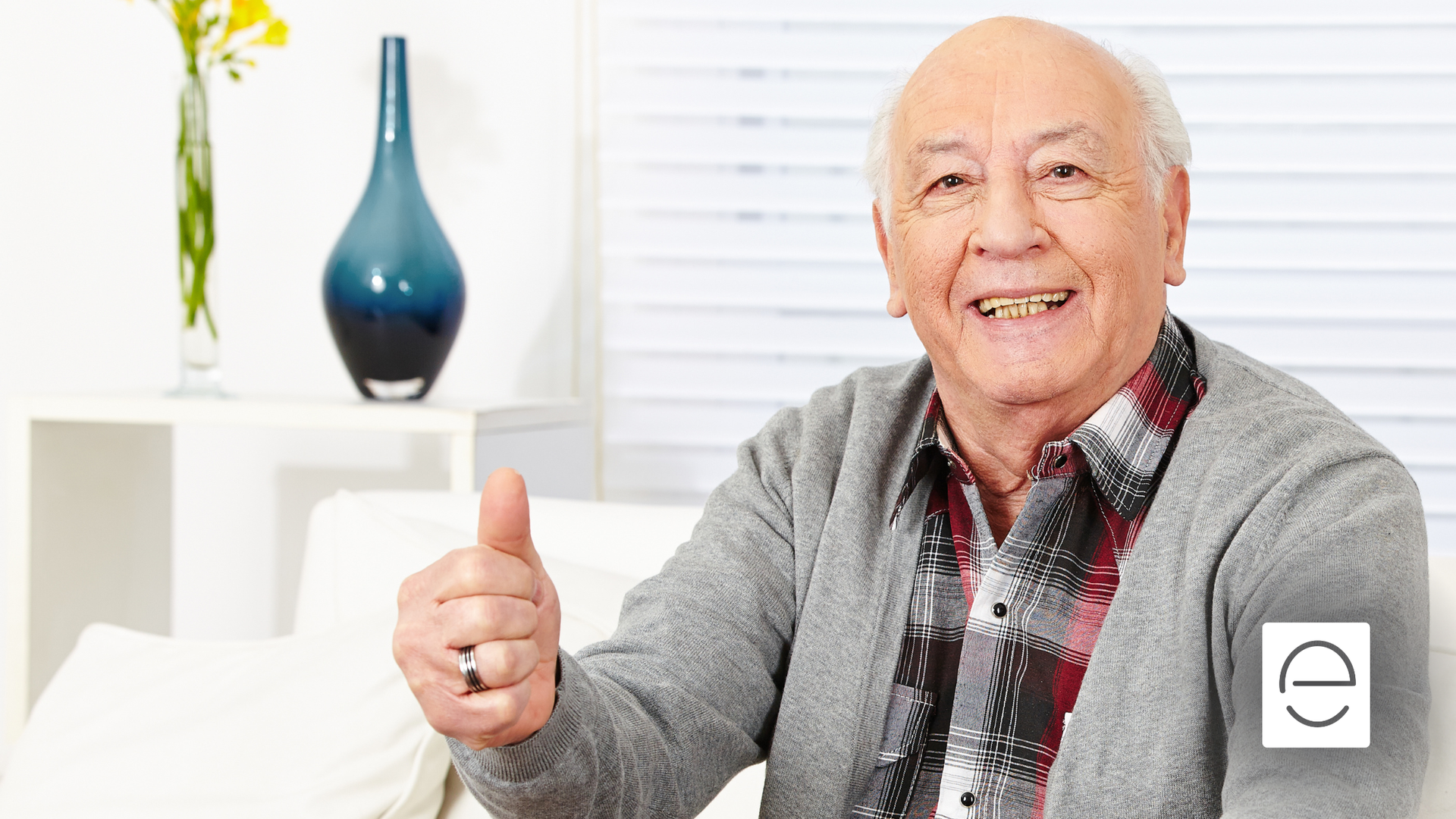 Older man in plaid shirt and gray sweater gives a thumbs-up, smiling on a white sofa.