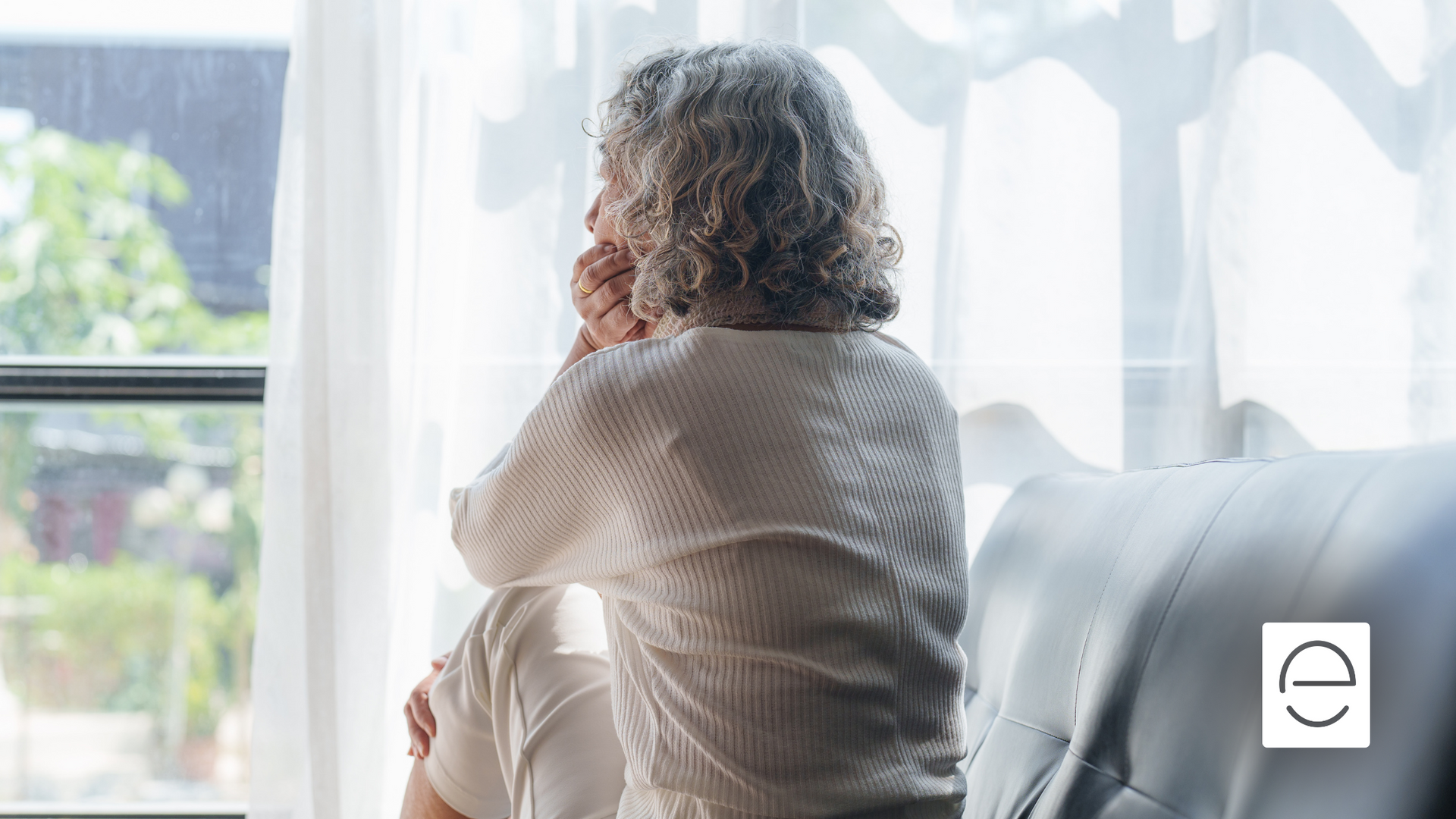 An elderly woman is sitting on a couch looking out of a window.