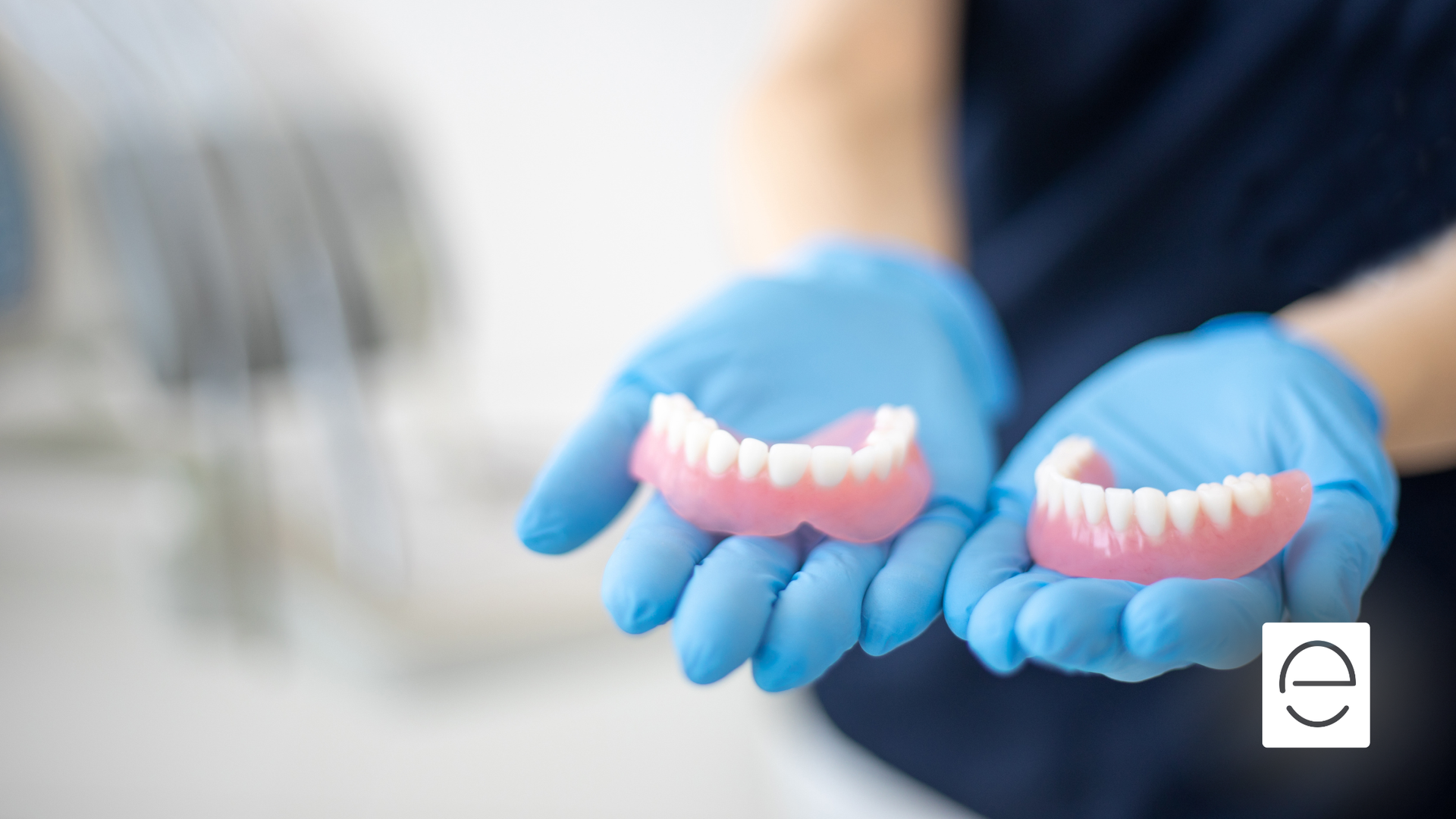 Hands in blue gloves holding two dentures against a blurred dental office backdrop.