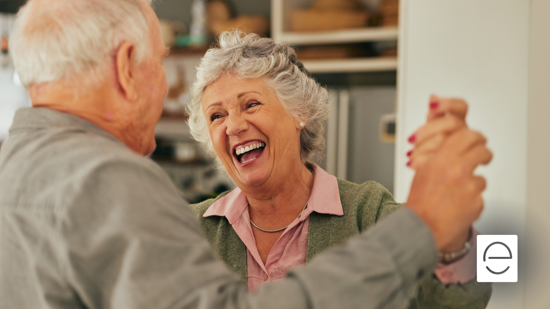 Elderly couple happily dancing indoors, woman laughing, warm lighting.