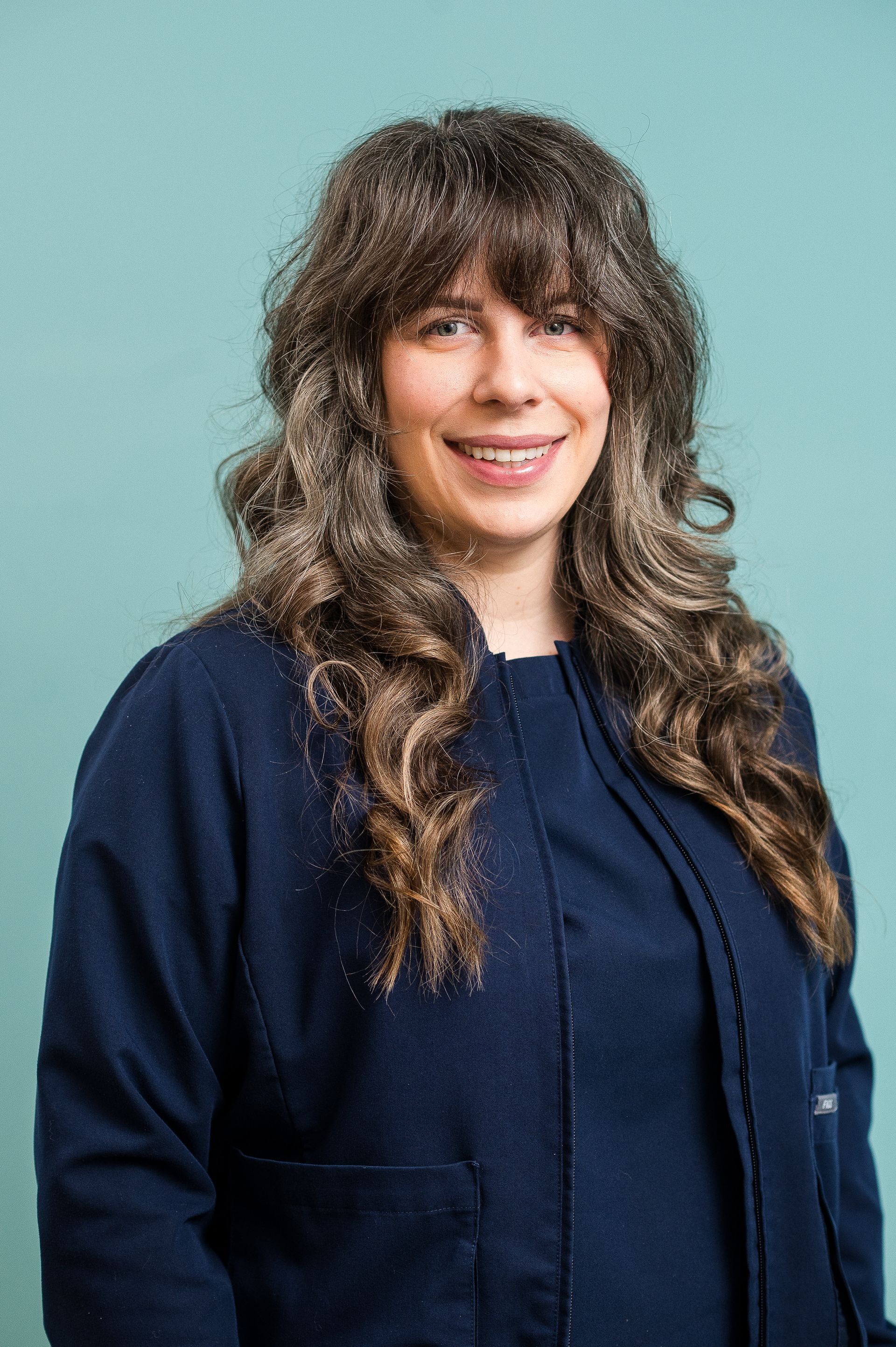 Woman with long curly hair smiling, wearing a navy jacket against a teal background