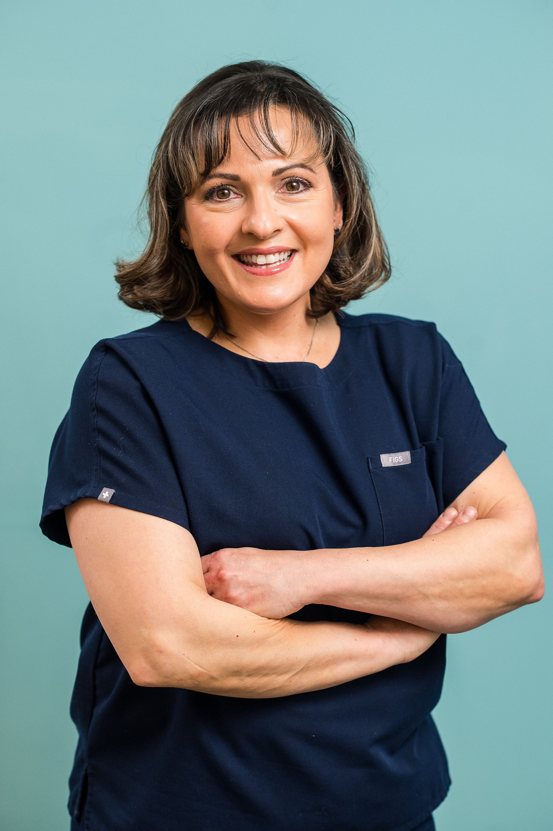 Smiling person in navy scrubs with arms crossed against a teal background