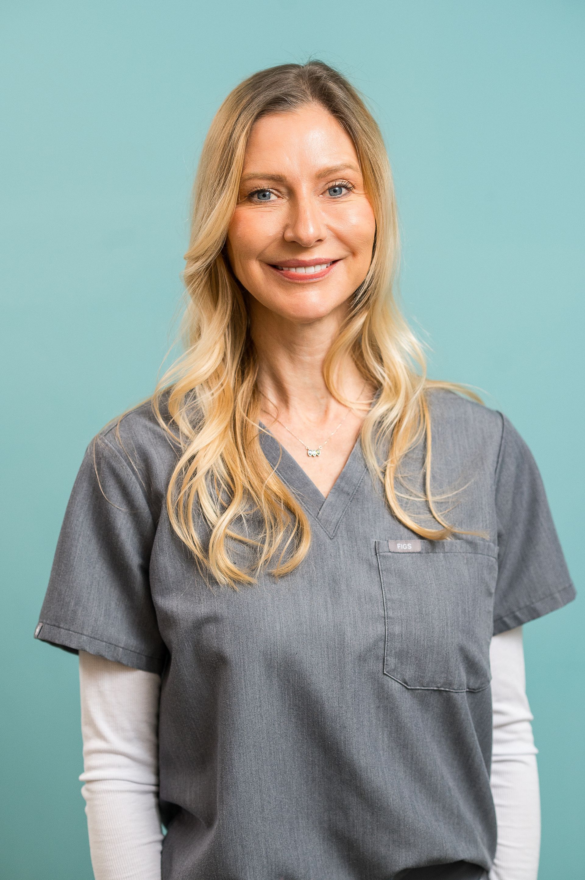 Smiling person in gray scrubs and white undershirt against a teal background