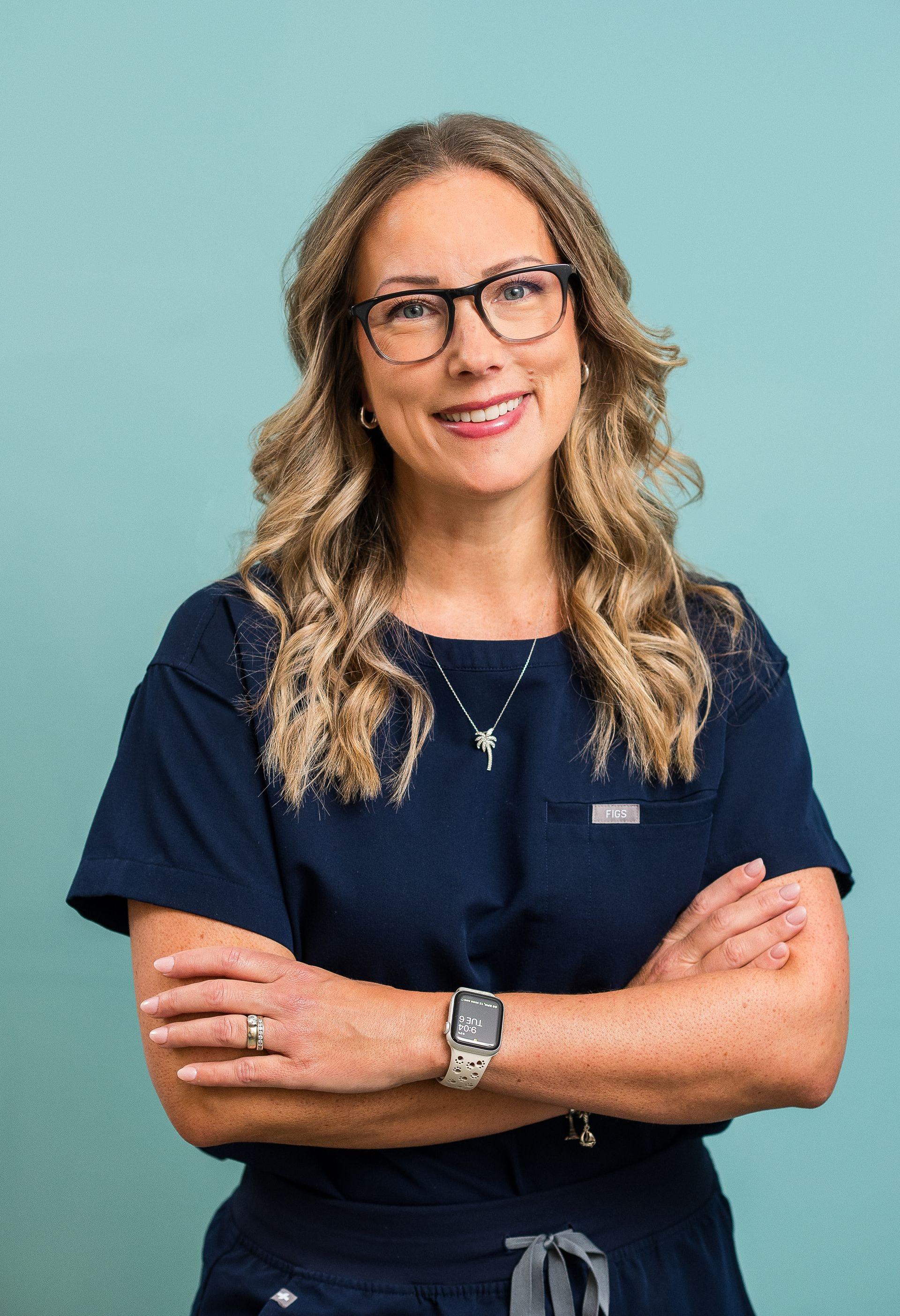 Smiling person in dark navy scrubs with arms crossed against a light teal background