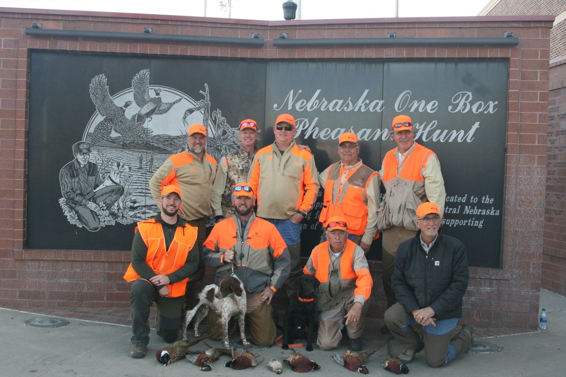 Group of hunters in orange vests pose with dogs and harvested pheasants at the Nebraska One Box Pheasant Hunt.