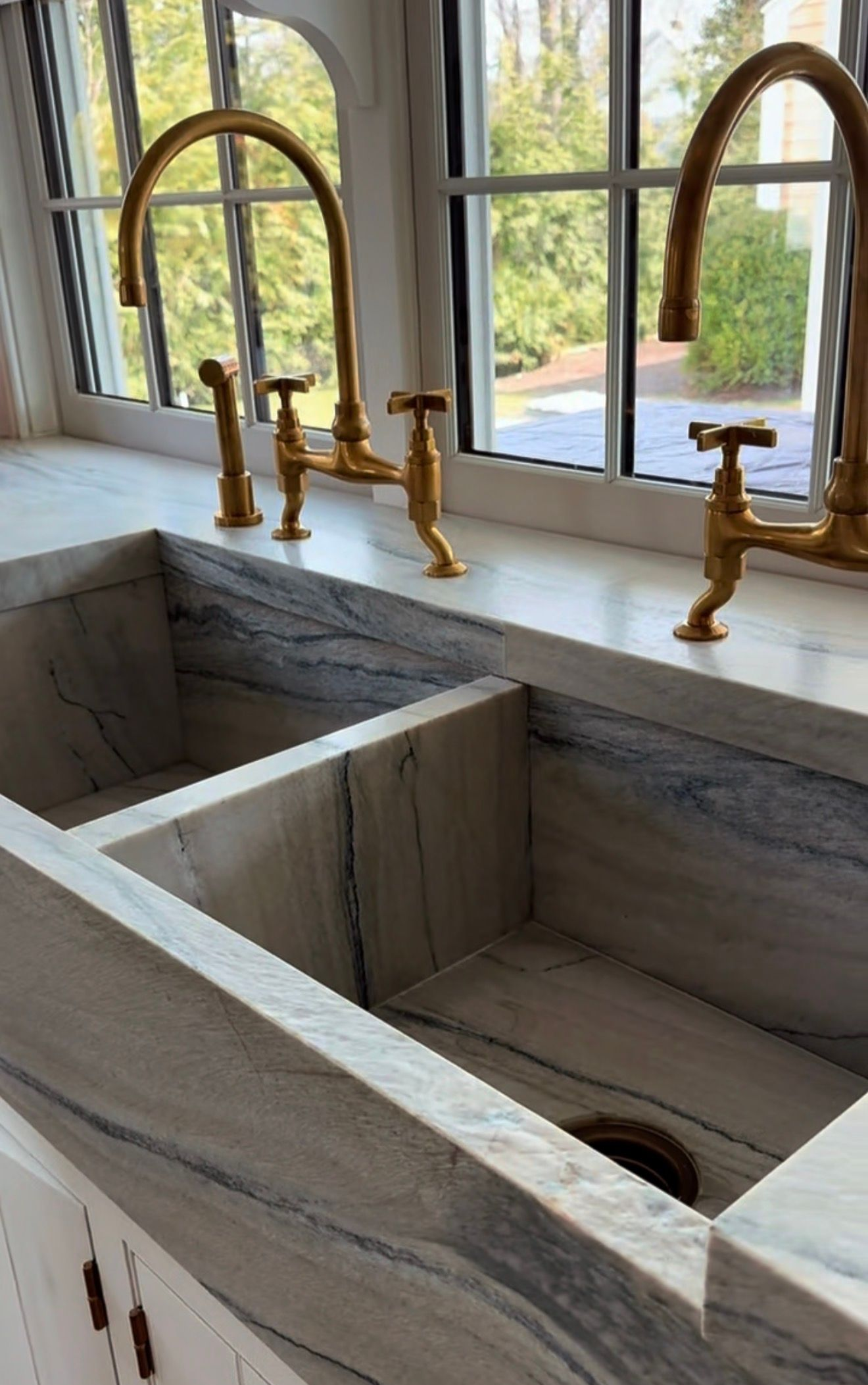 A marble, double-basin farmhouse sink with two brass bridge faucets sits beneath a window in a bright kitchen.