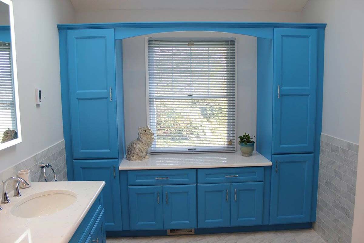 Blue bathroom cabinets surround a window with blinds; a white countertop and sink are nearby.