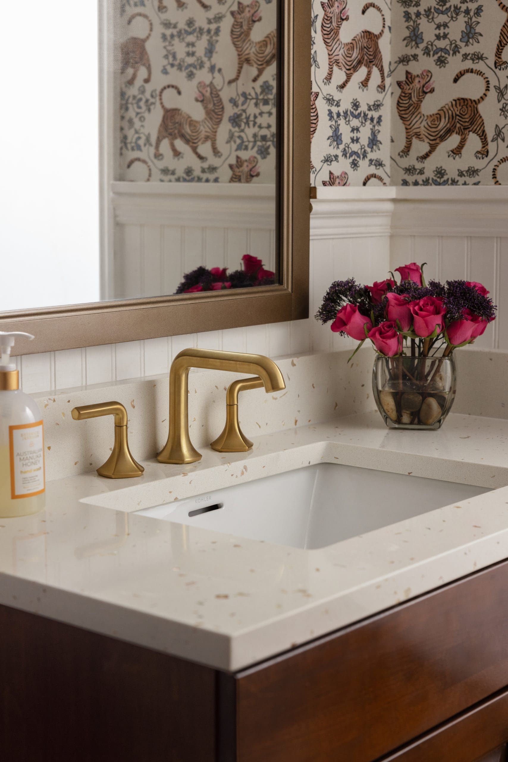 White bathroom vanity with gold faucet, mirror, and pink flowers in a vase.