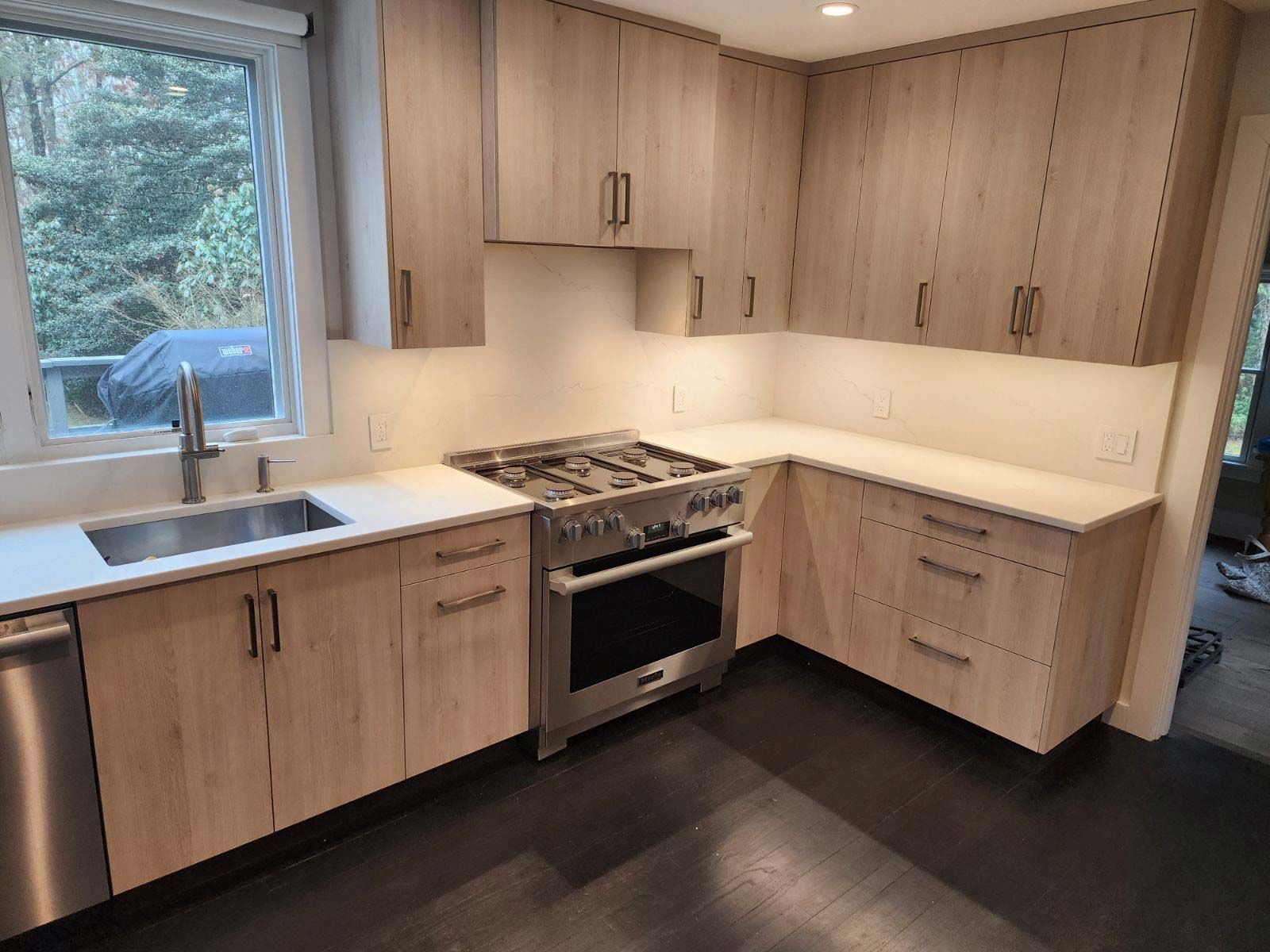 Modern kitchen with light wood cabinets, stainless steel appliances, and dark floors.