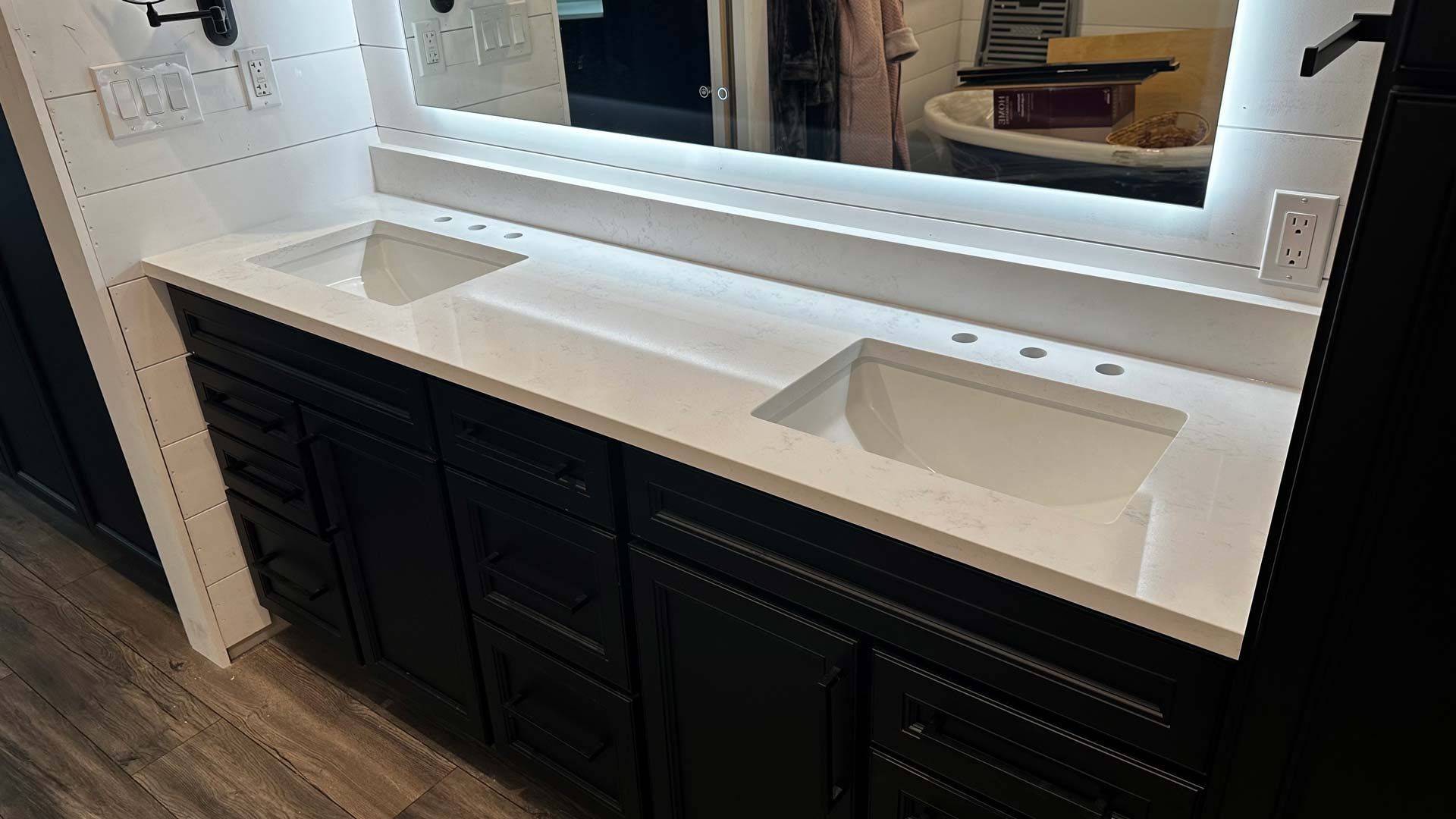 Bathroom vanity with white countertop, two sinks, black cabinets, and a lit mirror.