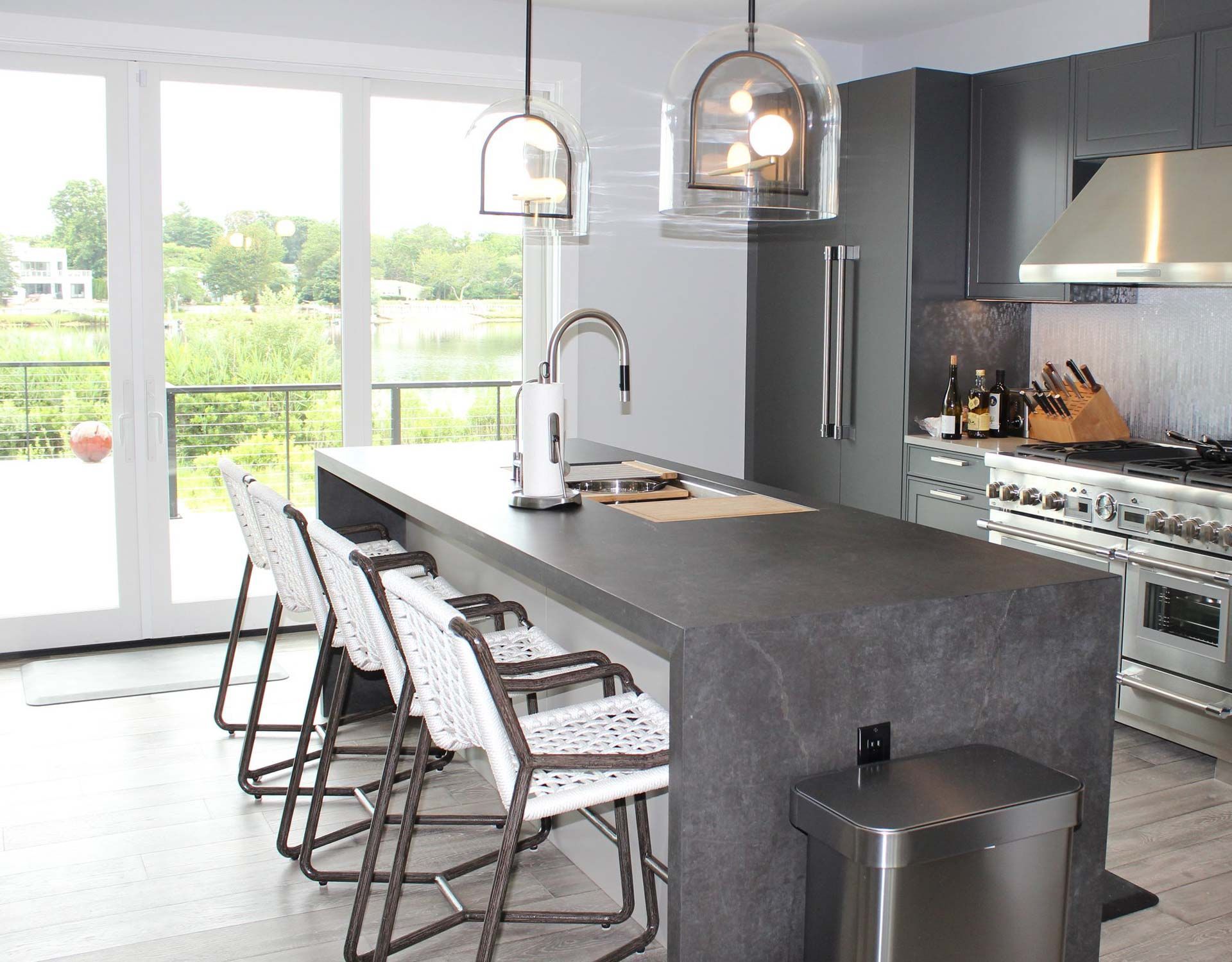 Modern kitchen with island, seating, and lake view. Dark gray cabinetry, stainless steel appliances, and white chairs.
