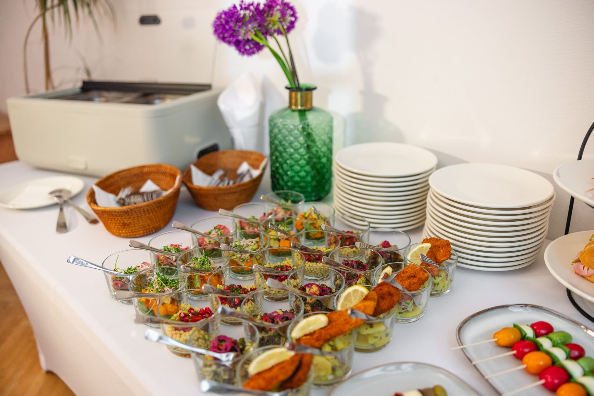 Buffet mit Vorspeisen, gestapelten Tellern, Weidenschalen und einer grünen Vase mit lila Blumen