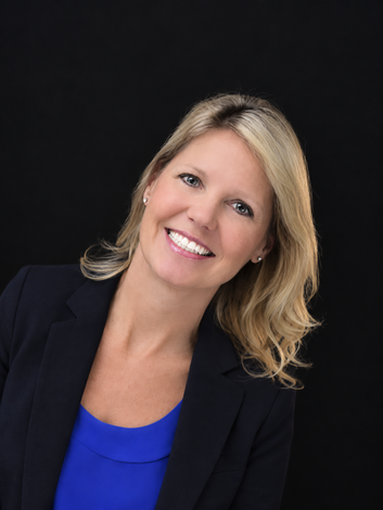 Smiling blonde woman in a black blazer and blue top against a dark background