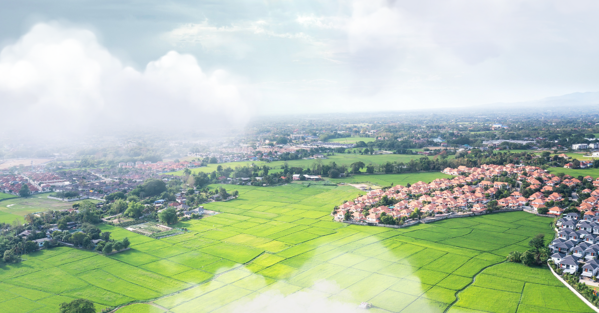 Aerial view: green fields, houses with brown roofs, cloudy sky.