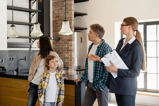Family and realtor viewing a kitchen. Blonde woman in suit points to space while the family observes.