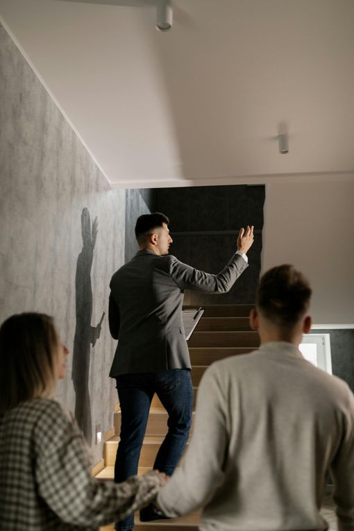 Real estate agent showing stairs to a couple, pointing up. Grey walls and light fixtures.