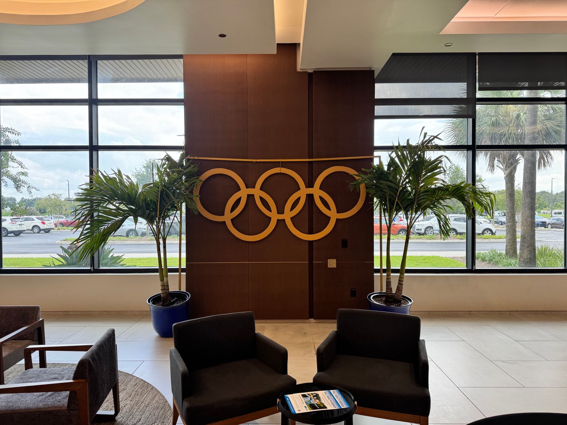  Indoor Olympic-themed decoration featuring gold rings mounted on a wooden panel, flanked by two potted palm trees,
