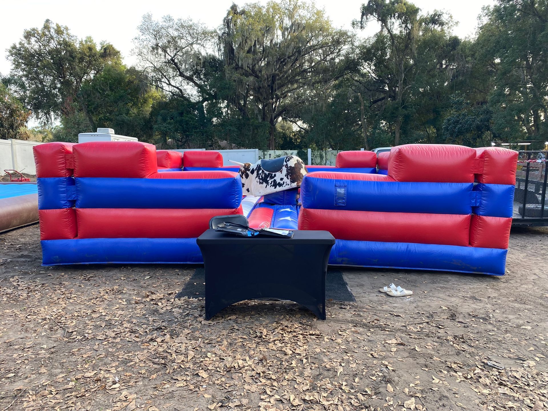 A mechanical bull ride setup outdoors on a dirt ground, surrounded by inflatable red and blue safety cushions. 