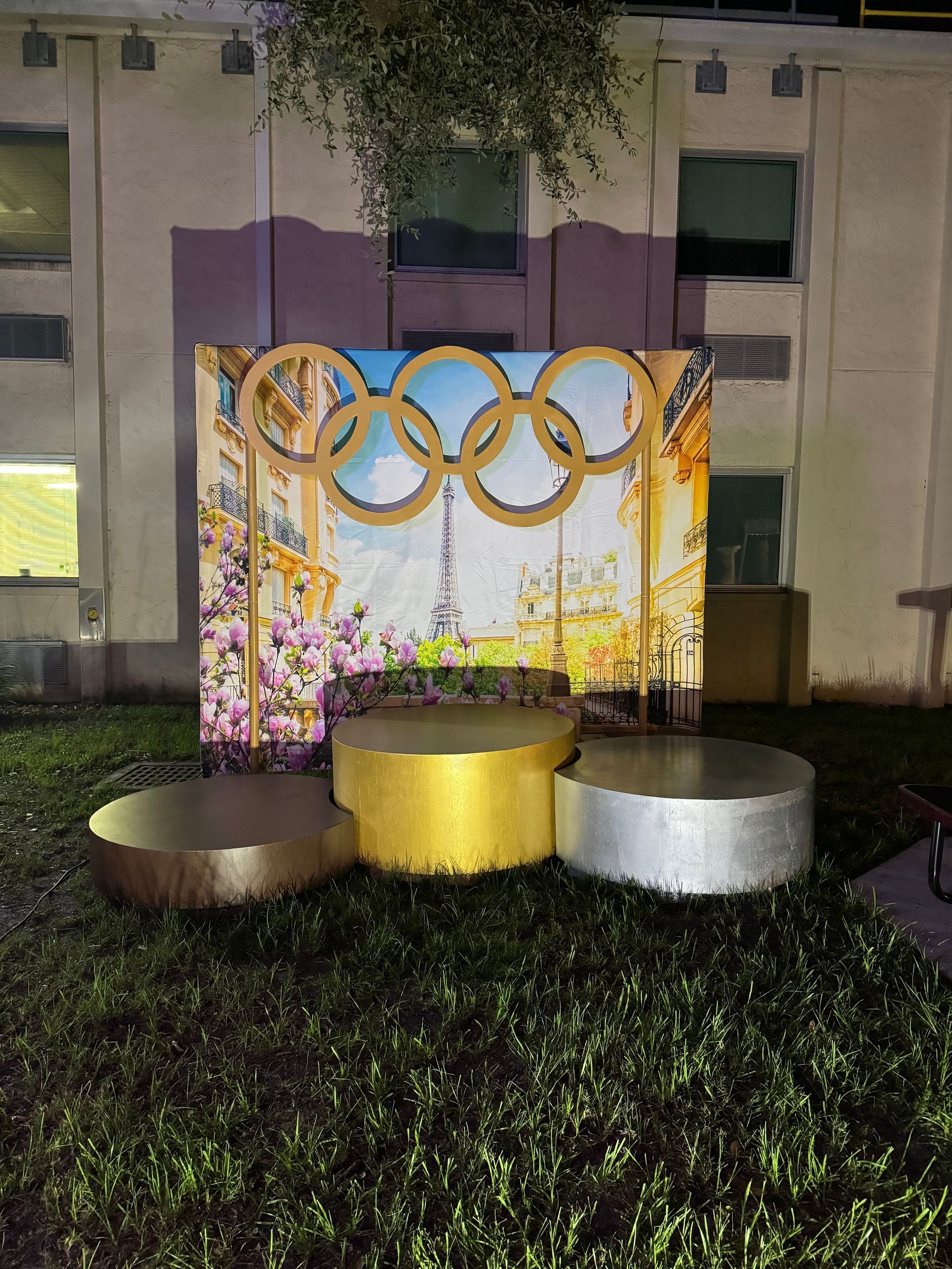  Paris Olympic-themed photo backdrop with gold rings, Eiffel Tower image, and a podium at night.