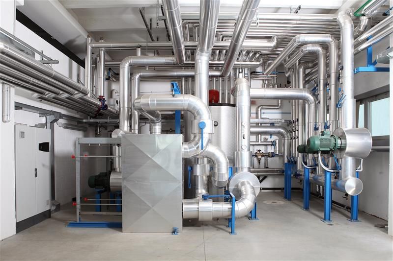 A bright, industrial mechanical room filled with complex, silver-insulated piping and a large central storage tank.