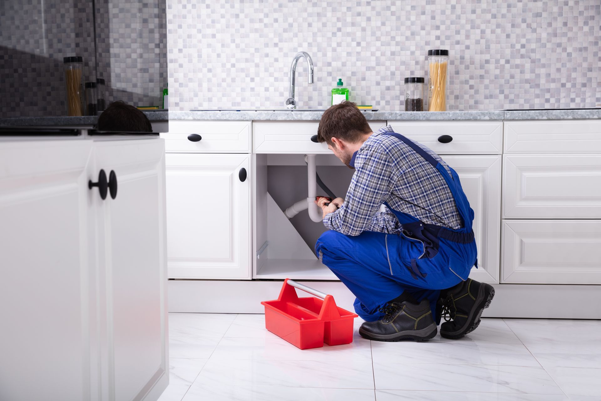 A plumber in blue overalls kneels on a white tile floor while repairing pipes under a kitchen sink.