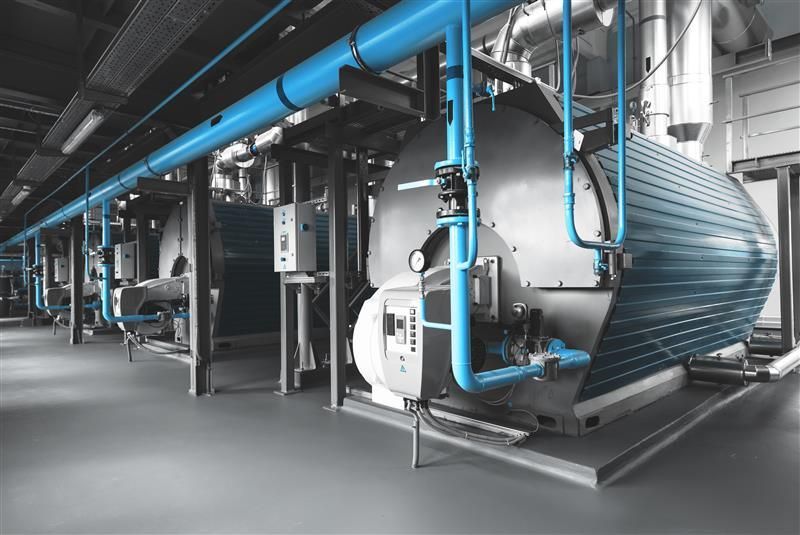 A row of industrial steam boilers in a facility, featuring large cylindrical tanks connected by bright blue pipes.