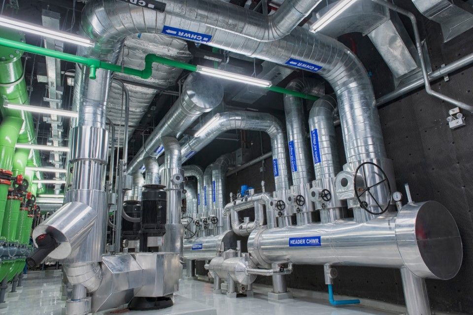 Industrial mechanical room with large, silver-insulated pipes and green conduits running along a dark ceiling.