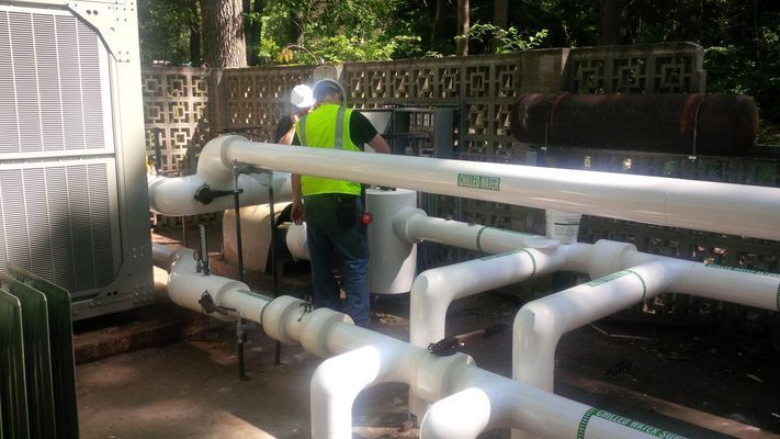 A worker in a high-visibility vest repairs white PVC plumbing pipes connected to a large outdoor unit.