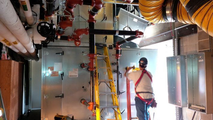 A person in a safety harness stands on a ladder working on large industrial machinery inside a utility room.