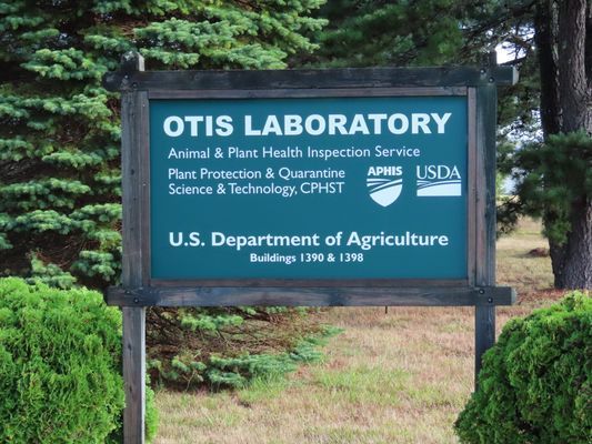 A sign for the USDA Otis Laboratory, located outdoors surrounded by pine trees and shrubs.