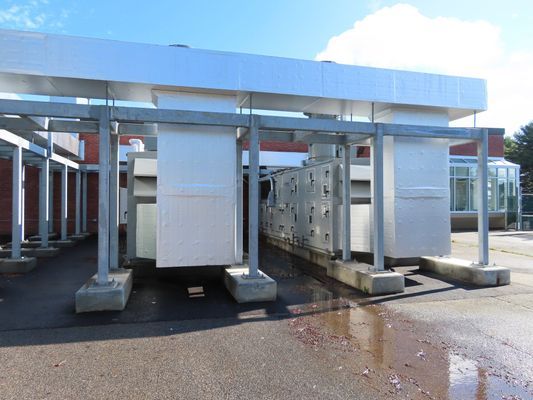 A row of large, white industrial HVAC air handling units installed on a concrete outdoor pad under a protective overhang.