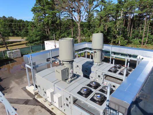 An industrial HVAC unit with two large, gray exhaust stacks positioned on a flat roof, set against a wooded background.