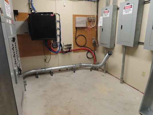 An electrical room featuring gray metal cabinets, conduits, and equipment mounted on a wooden backboard against white walls.