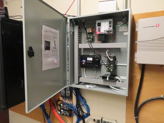 An open electrical equipment enclosure mounted on a wall containing wiring, controllers, and a small computer component.