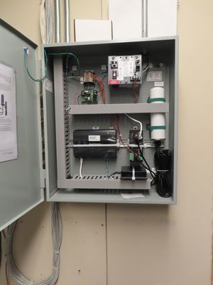 An open electrical control panel box mounted on a wall containing wires, modules, and a white cylindrical component.