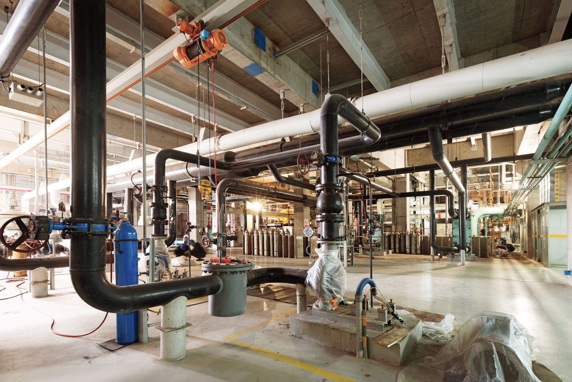 Industrial facility interior with extensive black and white piping, overhead equipment, and a concrete floor.