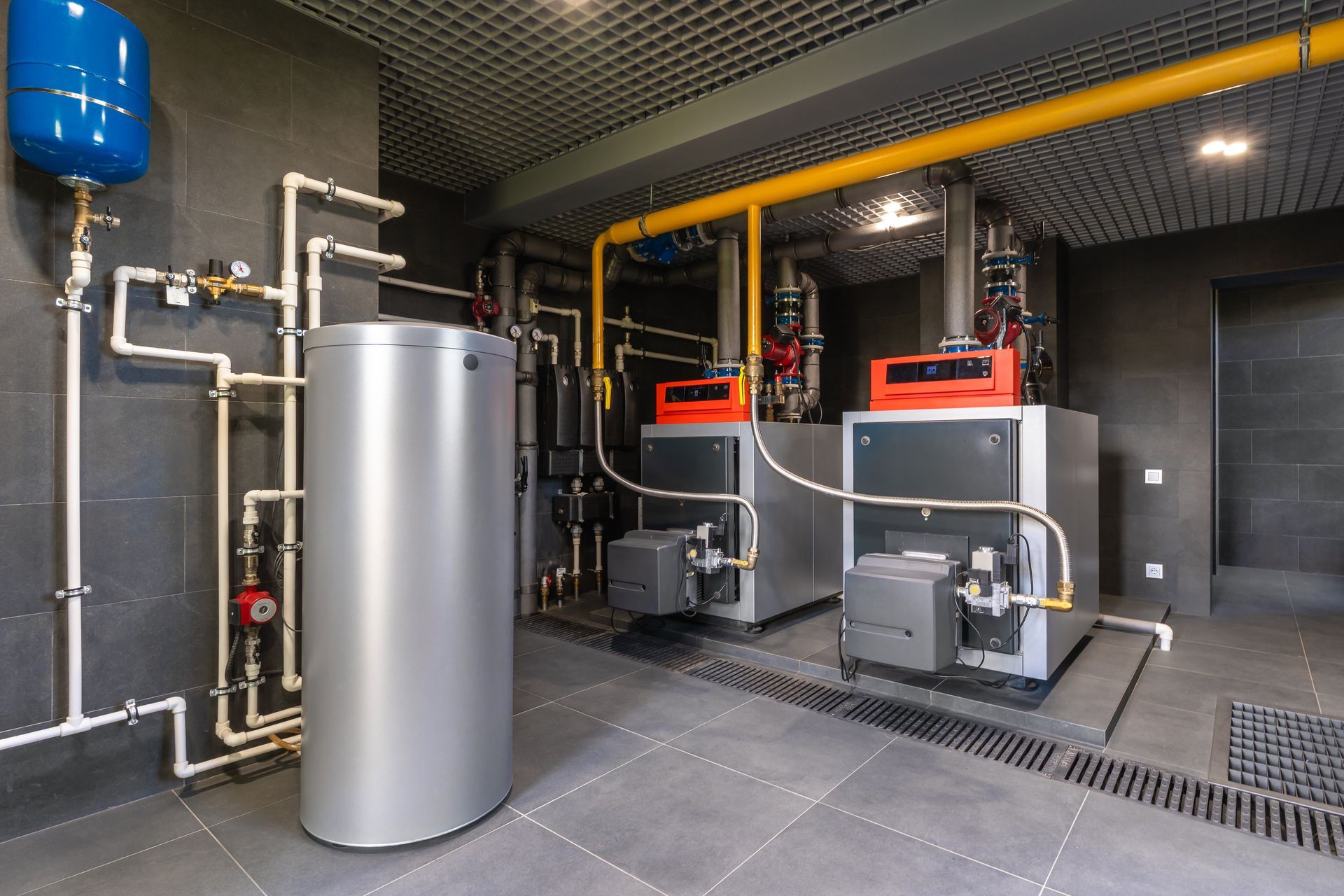 Two industrial boilers and a large metal water tank in a mechanical room with grey tiled floors and yellow ceiling pipes.