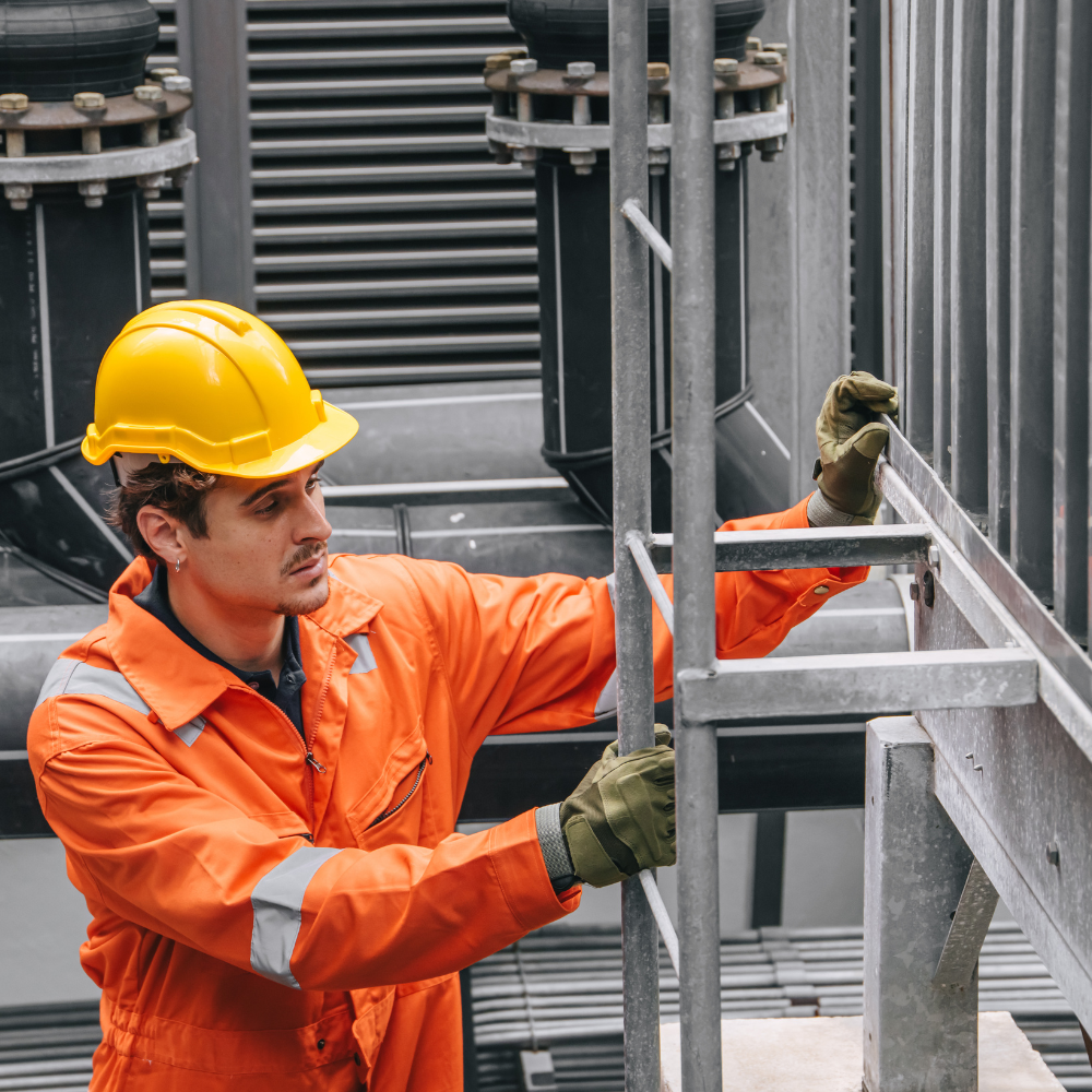 Worker in orange safety gear and yellow hard hat handling metal scaffolding at an industrial site.